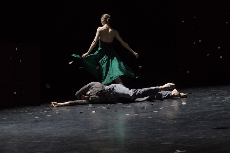A woman faces a way from the camera wearing a silk emerald green gown while a man lays on the floor.